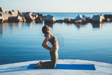 Foto de Vista lateral de la mujer concentrada en estera azul practicando yoga en rodilla con las manos en las caderas posición en el borde del terraplén - Imagen libre de derechos