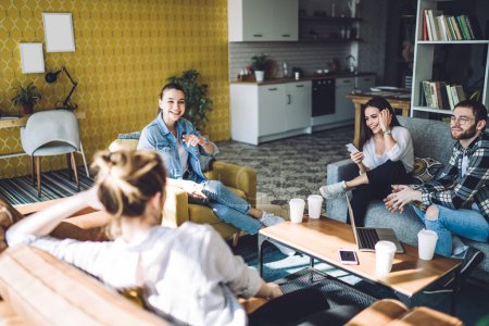 Téléchargez les photos : Jeunes étudiants de groupe de contenu se réunissant dans un appartement confortable boire du café avoir une conversation joyeuse tout en travaillant sur le projet avec un ordinateur portable - en image libre de droit