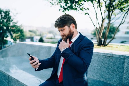 Exekutive fokussierten Mann in formeller Kleidung mit roter Krawatte mit Telefon und denken über Plan des Projekts sitzt auf Zaun im Park der modernen Stadt
