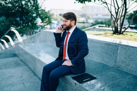 Empresario barbudo enfocado en traje con corbata roja discutiendo con el socio problemas de puesta en marcha utilizando móvil sentado en la cerca en el parque moderno de la ciudad