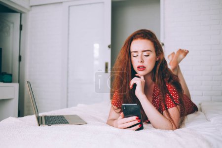 Photo for Pensive serious red-haired woman in bright clothing recreating on comfy bed while using smartphone and laptop in bedroom with trendy interior - Royalty Free Image
