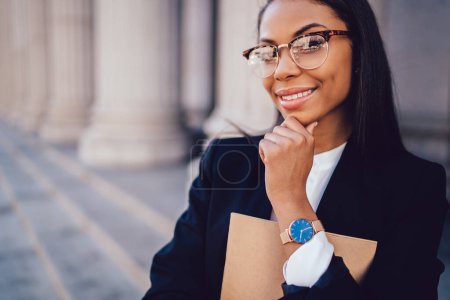 Emprendedora afroamericana positiva en ropa formal y gafas elegantes sonriendo y mirando a la cámara en la calle de Nueva York 