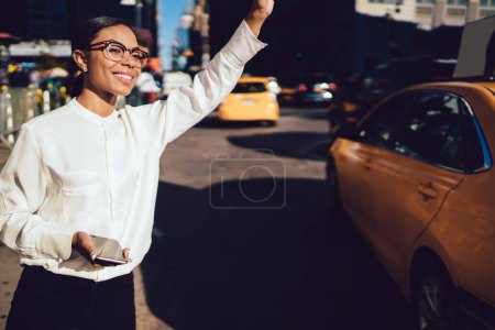 Gestionnaire afro-américaine de contenu en chemisier blanc à la mode et des lunettes élégantes souriant tout en levant la main pour taxi sur une journée ensoleillée à buste rue de New York 