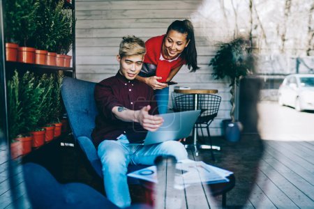 Photo for Smiling smart young multiracial coworkers in casual apparel talking with each other browsing laptop in creative workplace with potted plants - Royalty Free Image
