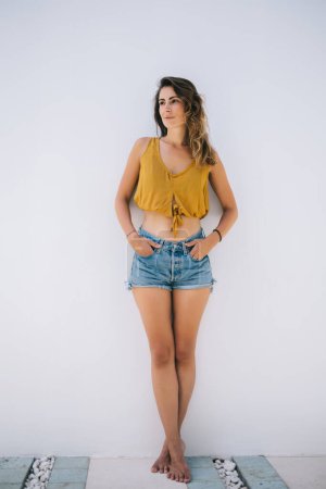 Photo for Thoughtful pensive romantic slender tanned shoeless woman in yellow top and jean shirts posing by wall and looking away on white background - Royalty Free Image