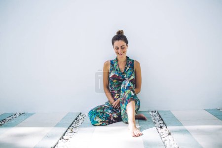 Photo for Satisfied modest bronzed woman in bright flashy apparel posing by wall while sitting cross-legged and looking down on white background - Royalty Free Image