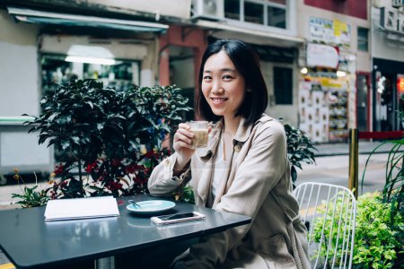 Photo for Serene positive ethnic Asian woman in casual wear sitting at table with copybook and smartphone while enjoying coffee on terrace in restaurant and looking at camera - Royalty Free Image