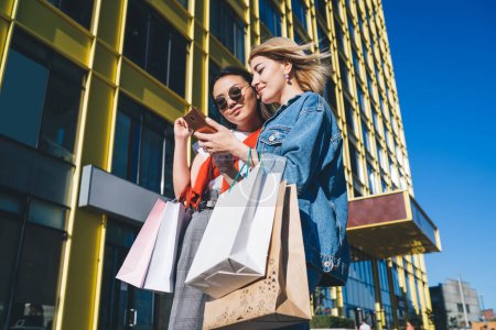Photo for From below smiling satisfied woman walking with ethnic lady in sun glasses with shopping bags using smartphone checking photos at outside of city mall - Royalty Free Image
