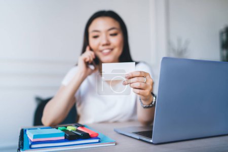 Jeune contenu intelligent femme asiatique en t-shirt blanc occasionnel parler appel sur téléphone mobile de travail avec ordinateur portable tenant la carte de visite dans la main dans le bureau