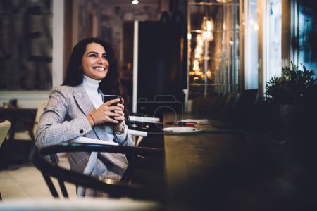 Kaukasische Teenagerin in lässiger Kleidung, die eine Tasse mit heißem Tee in der Hand hält und lächelt, während sie in der Freizeit in der Cafeteria denkt, glückliche Frau mit brünetten Haaren, die die freie Zeit zum Träumen im Café genießt