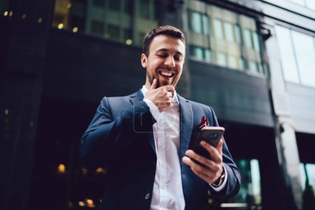 Optimistischer Manager im Anzug reibt sich das Kinn und lacht, während er auf der Straße steht und Nachrichten auf dem Smartphone in der Innenstadt der modernen Stadt liest