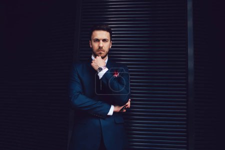 Thoughtful successful bearded businessman in suit looking at camera and touching chin with hand while standing at black striped wall