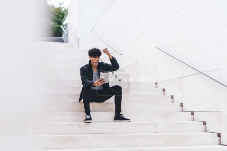 Téléchargez les photos : Angle bas de élégant heureux célébrant jeune homme afro-américain brunet en tenue décontractée navigation sur tablette tout en étant assis à l'escalier urbain sur la rue en plein jour - en image libre de droit