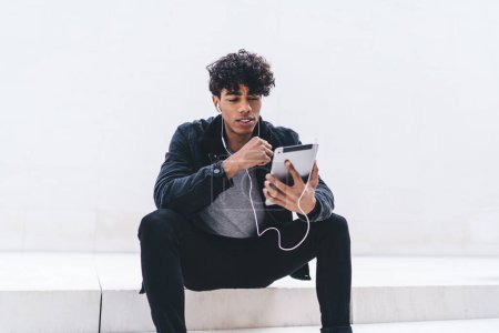 Stylish ethnic young man with earphones sitting on stairs with legs spread and looking at tablet while listening to music and checking playlist on white background