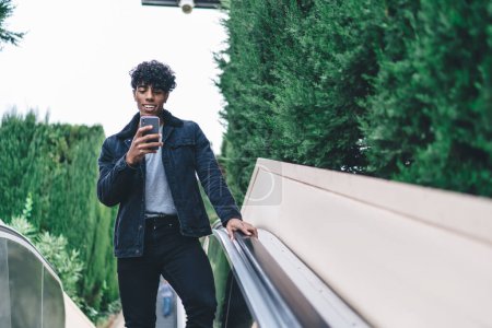 From below of happy handsome curly ethnic adult male in black jacket and jeans browsing smartphone on elevator on street on background green trees 