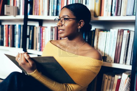 Photo for Black clever focused female sitting with book in library and leaning on bookshelf with literature during exam preparation looking away - Royalty Free Image