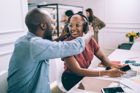 Photo for African American man putting on headphones to smiling stylish female colleague while working together with mobile phones and papers at workplace looking at each other - Royalty Free Image