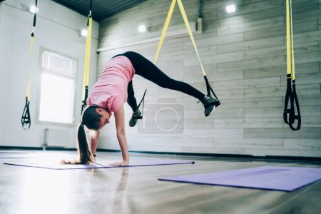 Attractive fit sportswoman wearing pink t-shirt and black leggings standing on hands while stretching legs using suspension belt in fitness studio