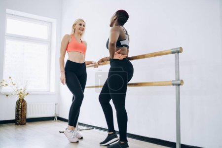 Smiling sportive confident multiethnic women in sportswear standing face to face in fitness center exercise room and talking while leaning on ballet barre