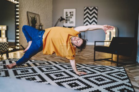 Foto de Hombre concentrado en ropa casual practicando yoga en alfombra haciendo meditación apoyada en la mano y la pierna en apartamento moderno - Imagen libre de derechos