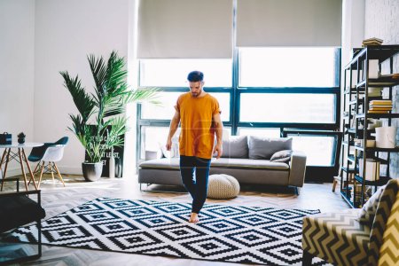 Téléchargez les photos : Jeune homme vêtu de t-shirt jaune et de jeans debout sur un tapis de yoga au milieu de la pièce et se préparant à faire des exercices de yoga - en image libre de droit