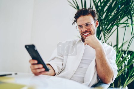 Smiling stylish bearded spectacled male dressed in white shirt browsing mobile phone while sitting at table with hand on chin in hall with indoor plant behind