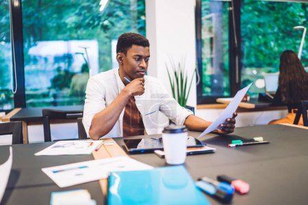 Hombre afroamericano frotando barbilla y reflexionando mientras está sentado en la mesa y leyendo papel durante el trabajo en el espacio de trabajo creativo moderno