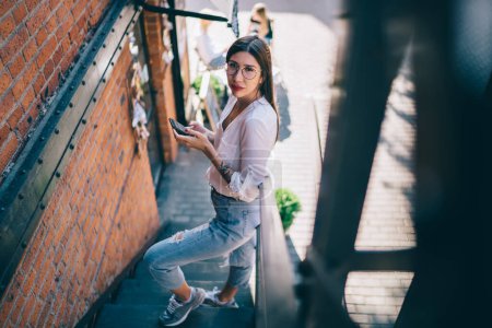 From above of Asian female in glasses wearing white blouse jeans and sneakers browsing mobile phone while standing on fire escape and looking at camera