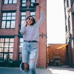 Positive hipster girl in joyful mood feeling happiness and excited during leisure time outdoors dancing from music podcast listen via electronic headphones and smartphone application for generation