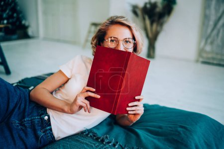 Pretty playful smiling young woman in casual outfit with glasses hiding behind book with red cover while lying down on bed in bedroom