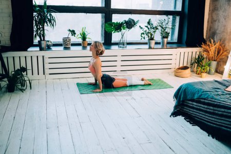 Sportliche junge Frau in stylischem Trainingsanzug, die morgens Stretchübungen für das Training macht, kaukasische gut aussehende Sportlerin, die in ihrem gemütlichen Zimmer in der eigenen Wohnung Yoga-Posen genießt