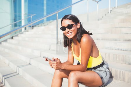 Portrait de blogueur joyeux dans des lunettes de soleil à la mode avec rebord de renard assis à l'urbanité de la ville touristique et en utilisant un gadget smartphone pour téléphoner en ligne, heureuse fille hipster latino bénéficiant de la mobilité