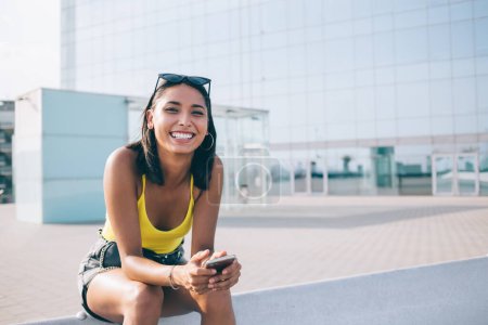 Portrait de dent réjouir fille hipster dans des vêtements élégants tenant gadget cellulaire moderne dans les mains et reposant sur le cadre urbain avec Internet public, blogueur femme prospère avec téléphone mobile