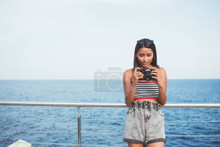 Front view of attractive millennial tourist checking photo images on modern technology standing near ocean during summer vacations, beautiful hipster girl editing pictures on advanced SLR camera