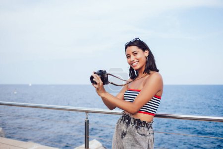 Portrait of smiling female tourist enjoying vacations time for recreation and leisure for creative hobby outdoors, smiling woman using SLR camera during promenade near ocean travel voyage