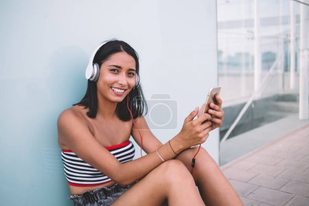 Téléchargez les photos : Portrait de gaie fille hipster dans les écouteurs pour l'annulation du bruit assis à l'urbanité et rire pendant le temps libre pour écouter de la musique positive playlist en utilisant smartphone application médiale - en image libre de droit