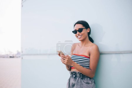 Demi-longueur du millénaire hispanique prospère avec un sourire sincère sur le réseau de visage sur gadget mobile pendant les loisirs à l'urbanité, fille hipster heureux dans les lunettes de soleil profiter du temps pour téléphoner en ligne