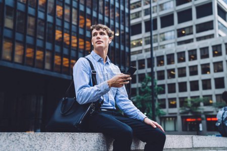 Jeune homme en chemise bleue et pantalon avec sac en cuir messagerie sur téléphone assis sur fond de New York