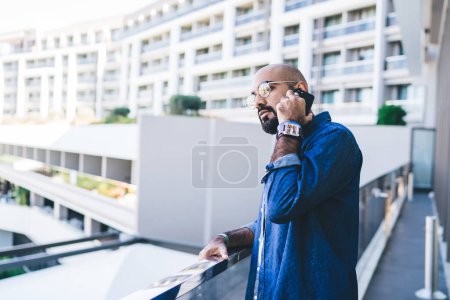 Photo for Side view of mature bald man in eyeglasses standing on balcony and talking via cellphone while looking away with pensive face - Royalty Free Image