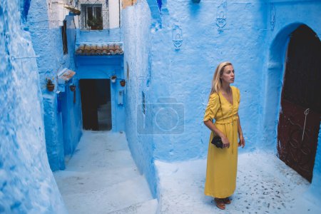 Von oben eine blonde Frau in gelbem Kleid mit Fotokamera auf der Schulter, die die Architektur der alten Stadt in Marokko bewundert, während sie neben einem alten Gebäude aus blauem Stein steht