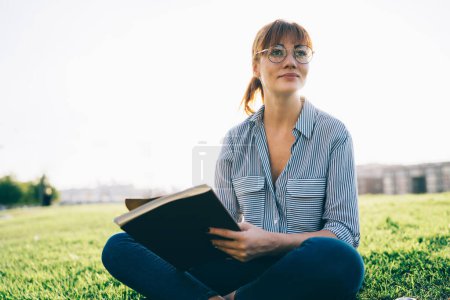 Attractive female author in eyewear pondering on article recreating in park on vacations, clever girl student doing homework writing in notepad looking away through eyewear for vision correction  