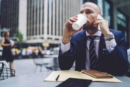 Foto de Hombre de moda vistiendo traje de negocios y corbata bebiendo de taza desechable y contestando teléfono sentado al aire libre con documentos y mirando hacia otro lado - Imagen libre de derechos