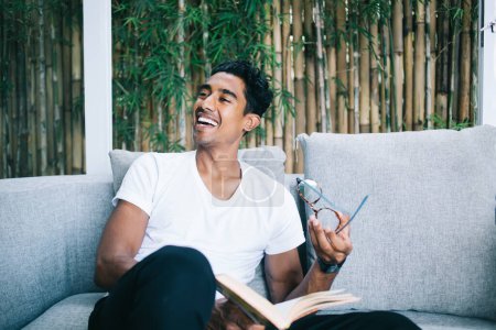 Happy young male with glasses and book in hands laughing while sitting on comfortable couch at home