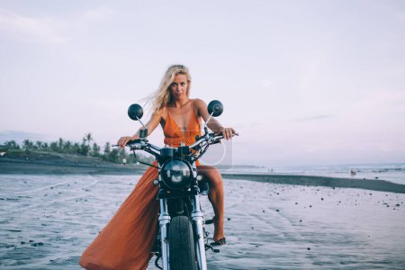 Photo for Attractive young female with long hair in orange dress sitting on motorcycle and looking at camera on beach at dusk - Royalty Free Image