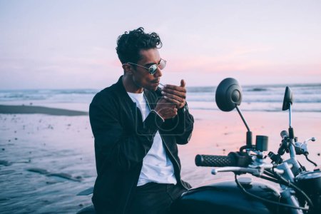 Élégant bel homme hispanique en lunettes éblouissantes et veste sombre éclaircissant cigarette et protégeant le feu contre le vent parking sur la moto en bord de mer le soir du coucher du soleil  