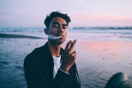 Photo for Tranquil Hispanic man in dark jacket holding cigarette and thoughtfully exhaling smoke and looking at camera in seaside on sunset evening - Royalty Free Image