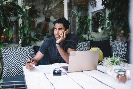 Nachdenklicher junger hispanischer Mann mit Stift in der Hand sitzt mit Laptop-Smartphone am Tisch und hält Papier in der Hand, während er im Restaurant kontempliert
