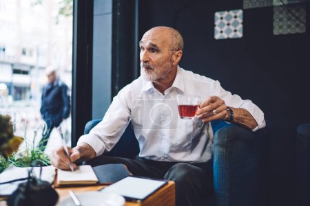 Photo for Old bald male in white shirt and beard and pen siting at table drinking tea while writing in notepad and using tablet in cafe looking away - Royalty Free Image