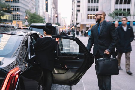 Confident polite African American businessman in suit opening car door for female colleague while standing on pavement in urban street