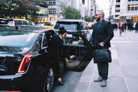 Erfolgreicher Manager mit schwarzer Glatze hilft weiblichen Mitarbeitern und öffnet Autotür, während sie auf dem Bürgersteig in der Stadtstraße steht